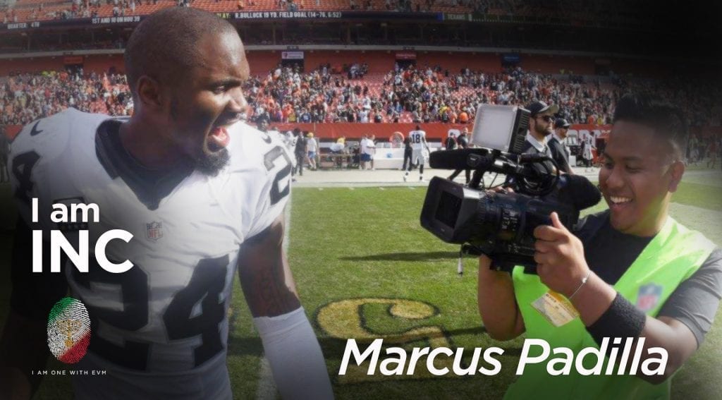 Man on a football field, holding a video camera pointed at a professional football player.