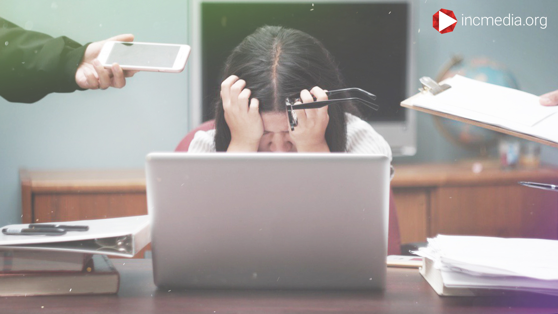Woman sitting at a desk in front of her laptop with her hands on her head while holding eyeglasses and people handing her a cell phone and clipboard.