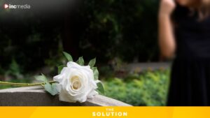 A person stands in front of a headstone with a large white rose laid on top of it
