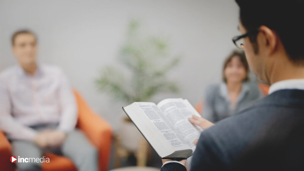A man in a suit reads from an open Bible to two seated individuals in a Bible study setting with the INC Media in the lower left corner.